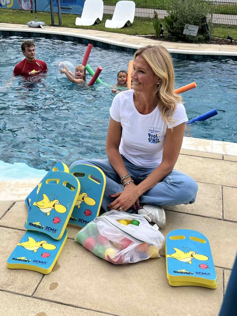 Franziska van Almsick sitzt im Schneidersitz am Poolrand. Hinter ihr schwimmen Kinder im Pool.
