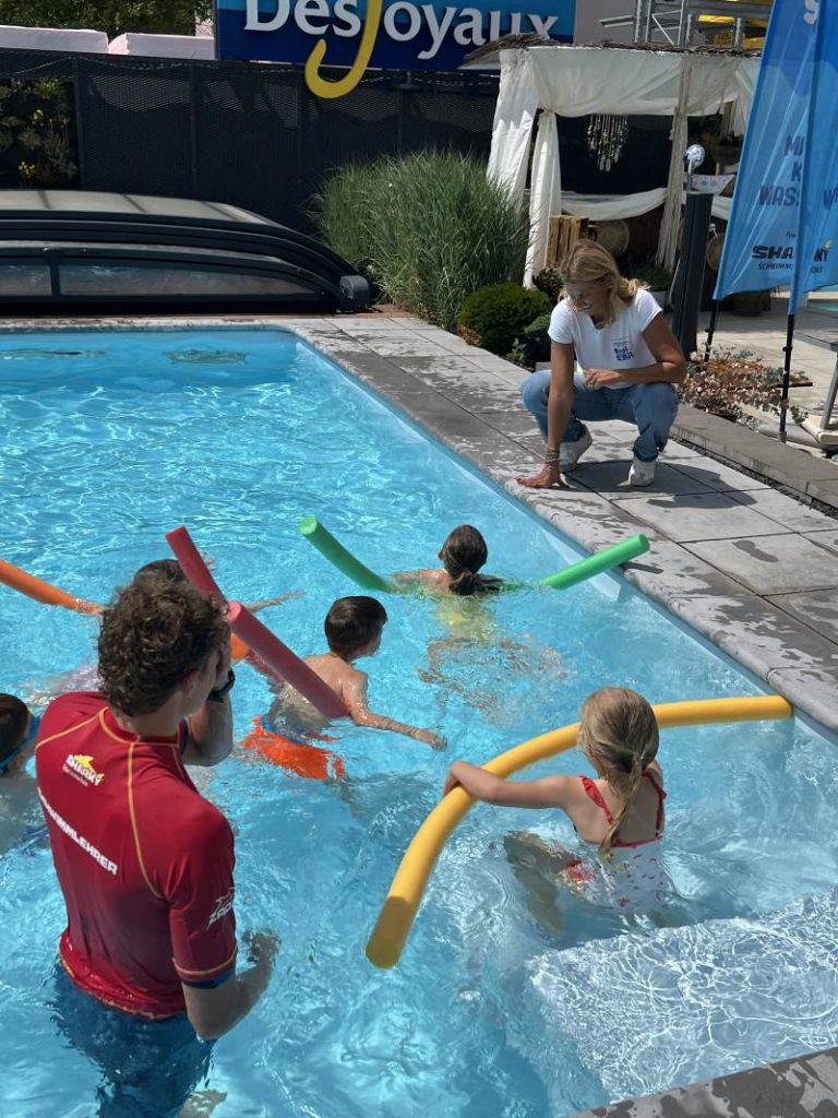 Kinder schwimmen im Pool. Der Schwimmlehrer begleitet sie. Franziska van Almsick schaut ihnen von Beckenrand aus zu.