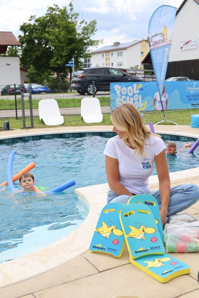 Franziska van Almsick sitzt im Schneidersitz am Pool. Sie schaut über die Schulter zu einem Kind im Wasser.