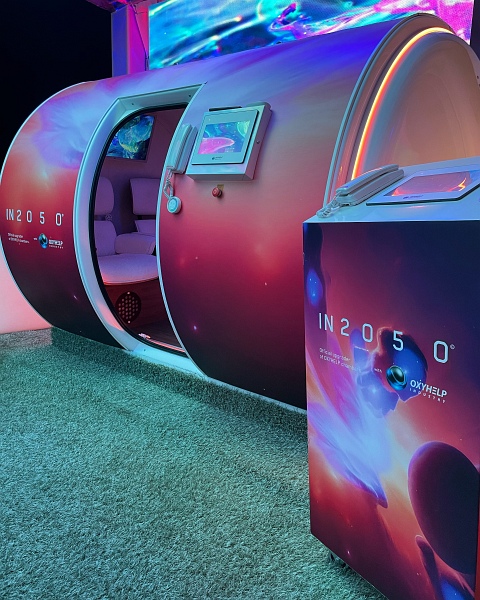 Zu sehen ist eine runde Kabine, deren Tür geöffnet ist. Die Tür ist geöffnet und im Innenraum sieht man 2 Sitze. Das Bild wirkt futuristisch an.