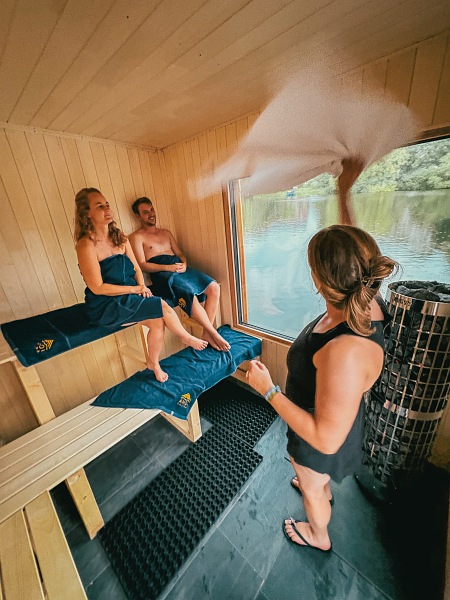 Zwei Besucher sitzen auf einer Bank in der Sauna. Eine weitere Person steht davor und wedelt mit dem Handtuch um den Dampf zu verteilen.