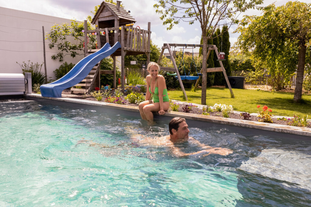 Eine Frau sitzt am Poolrand. Ein Mann schwimmt in dem Pool. Im Hintergrund ist ein Garten zu sehen.