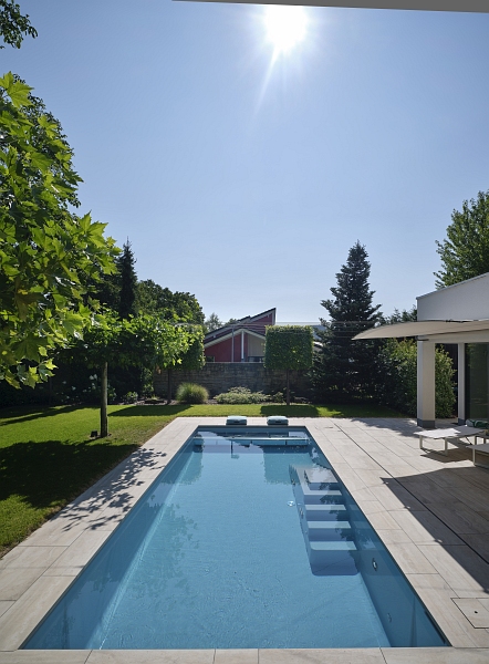 Der Pool wird von hellen Terrassensteinen umrandet und liegt im grünen Garten.